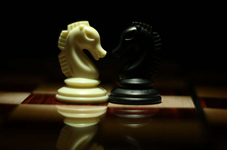 Black and white knight pieces from chess set sitting on a chess board under a spotlight.